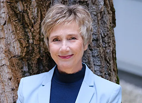 Amelie Fried lächelt in die Kamera, trägt einen hellblauen Blazer und dunkelblaues Oberteil vor einem Baumstamm.